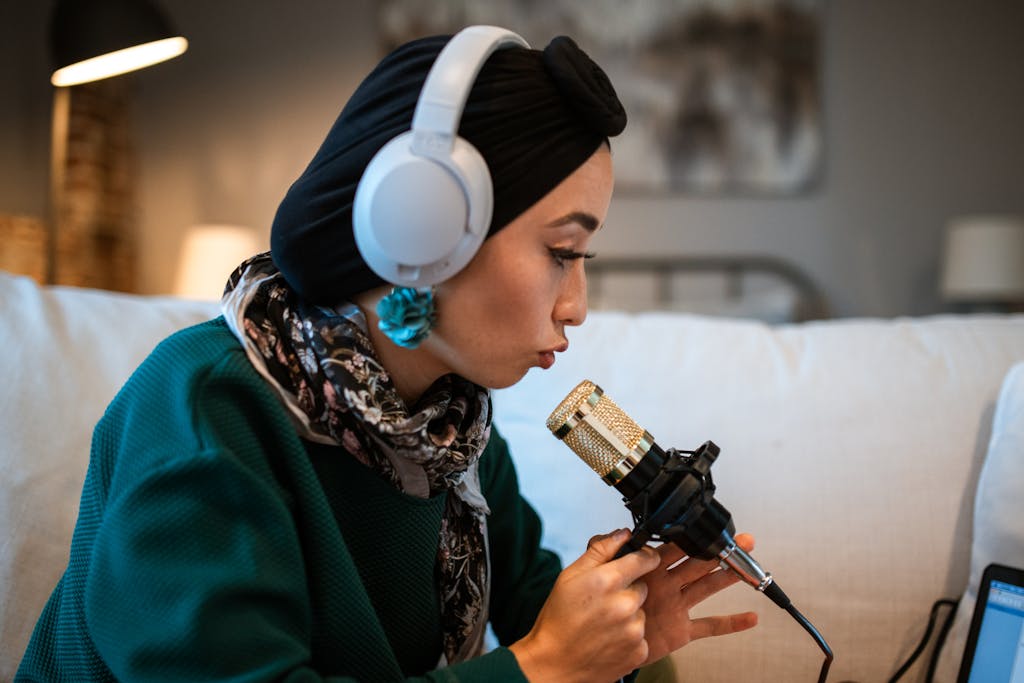 A woman wearing headphones records a podcast using a gold microphone indoors.