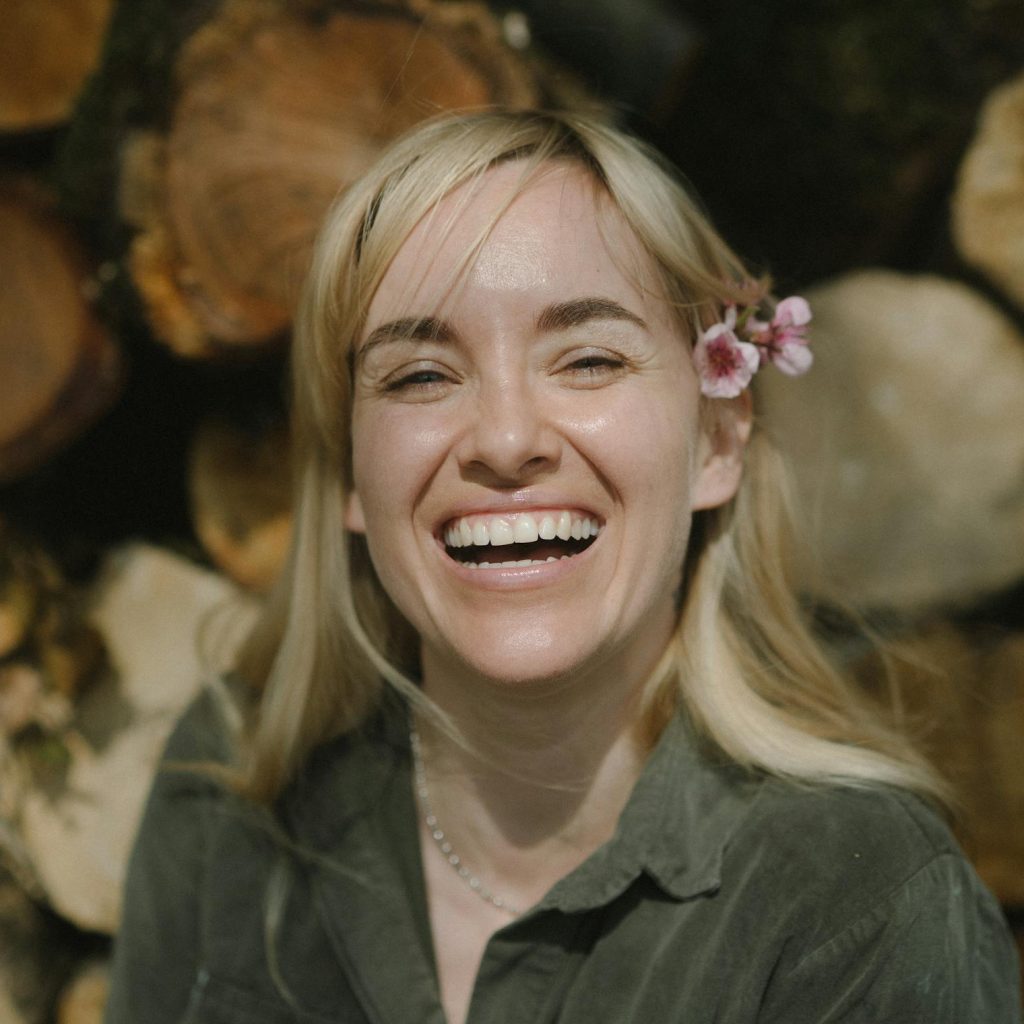Happy woman with cherry blossom in hair smiling outdoors against wooden logs.